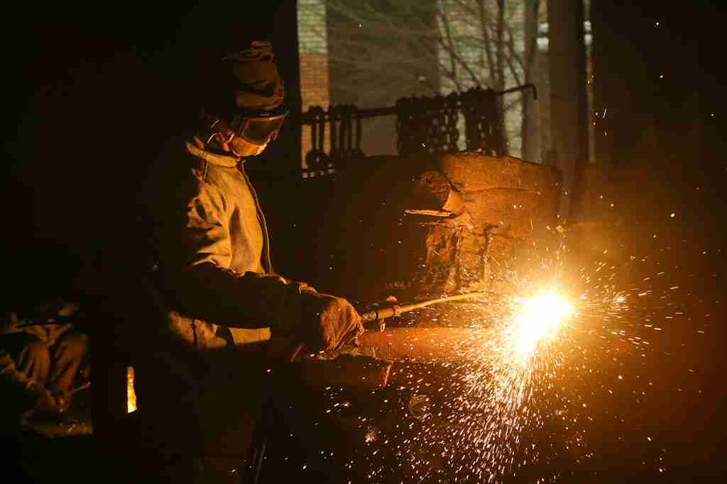 A welder in protective gear working on metal in a dimly lit workshop with sparks flying.